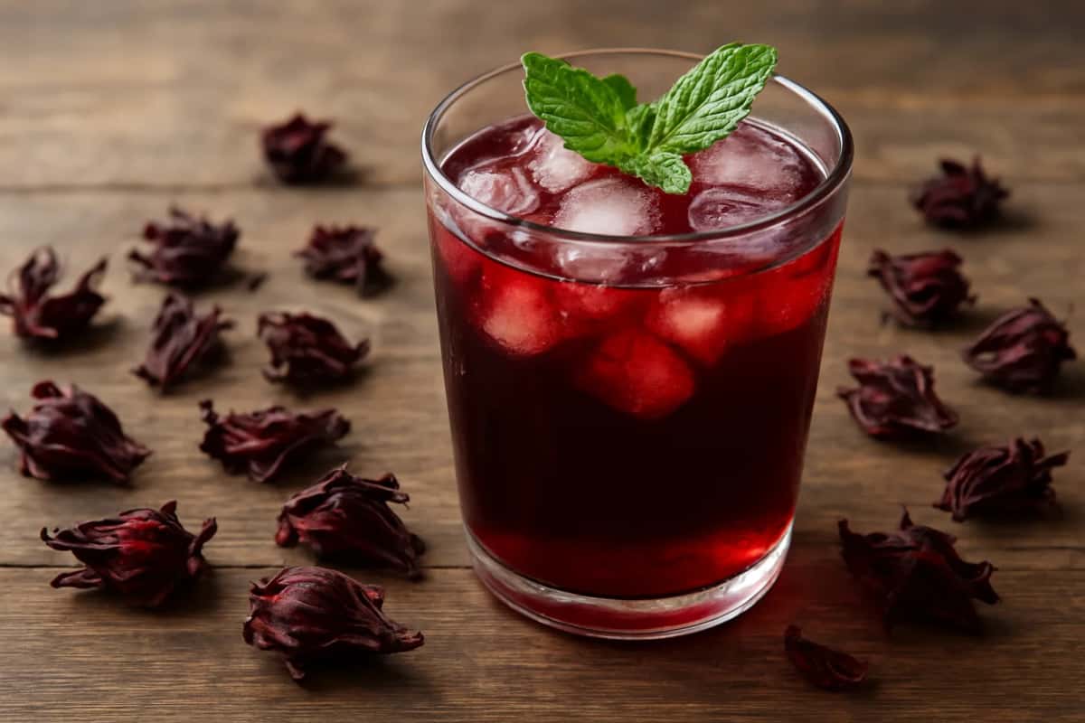 Verre de jus de bissap rouge foncé avec glaçons et feuilles de menthe, entouré de calices d'hibiscus séchés