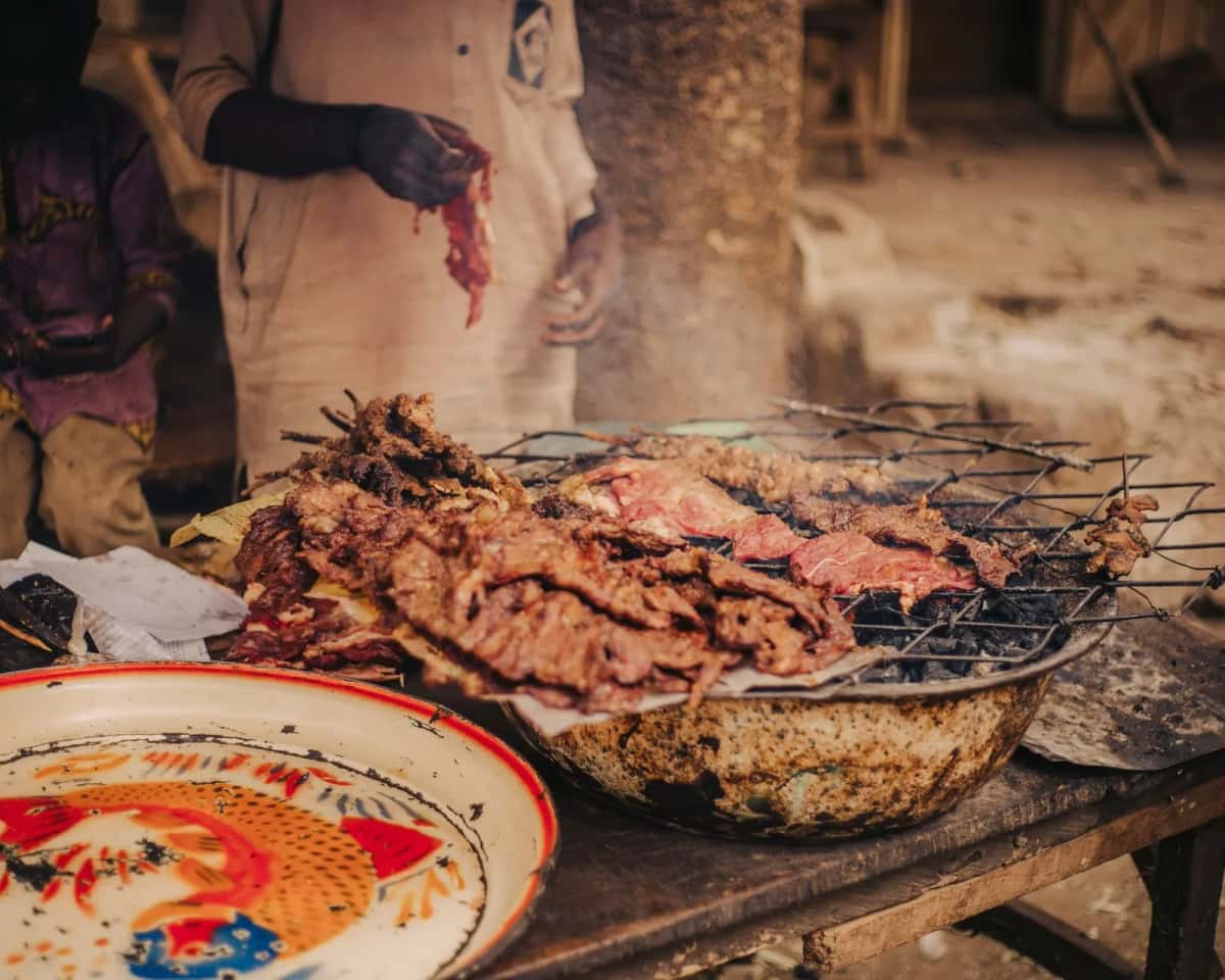 Viande grillant sur une grille posée au-dessus des braises, dans une échoppe de rue ouest-africaine, un grilleur debout à côté