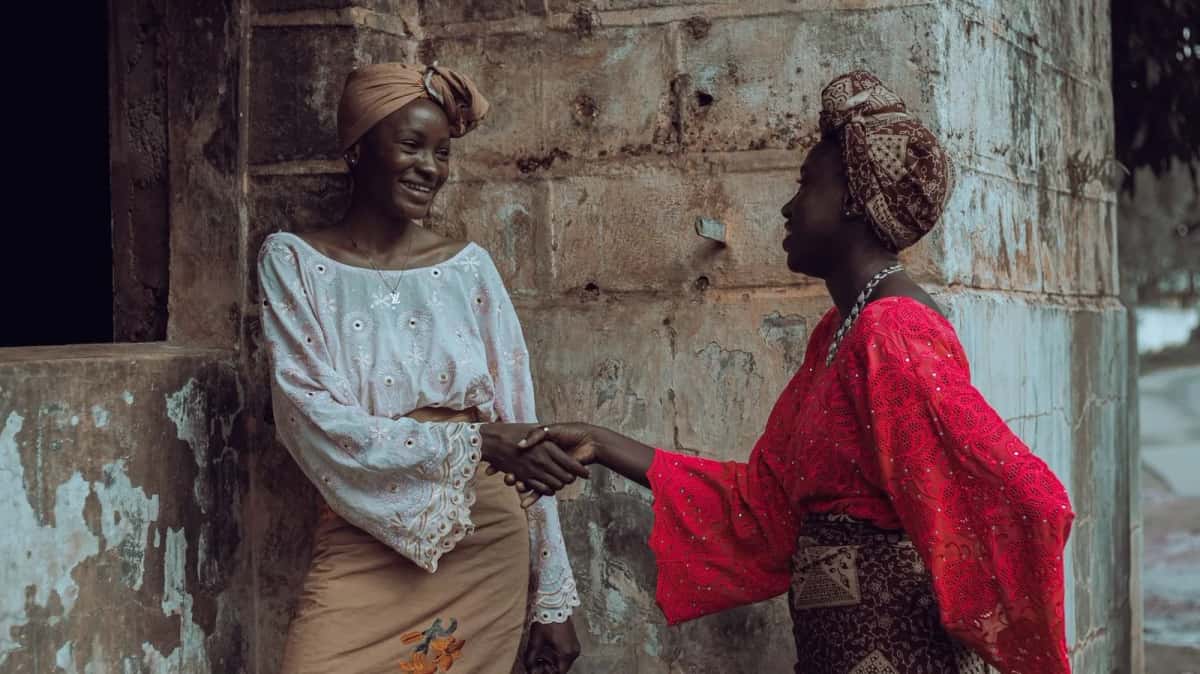 Deux femmes en foulards colorés se serrent la main en souriant devant un mur ancien
