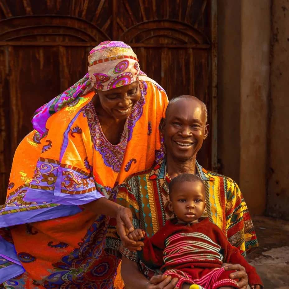 Grand-mère en boubou orange et grand-père en chemise wax, souriants, avec un bébé sur les genoux